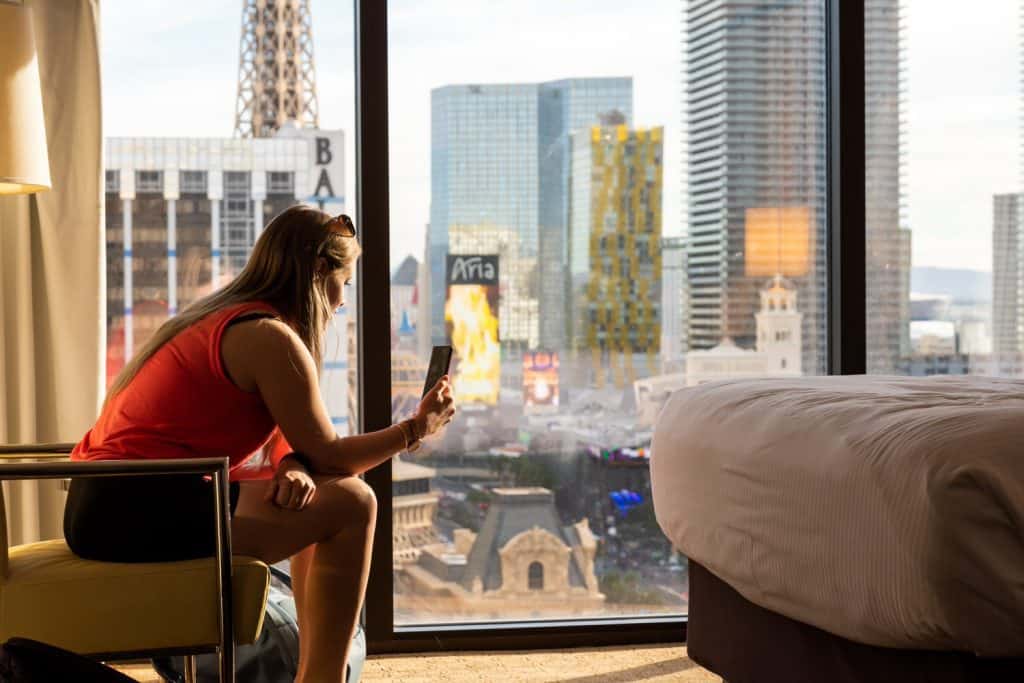 Girl sits on a chair on her phone in a hotel room with Las Vegas out the window.
