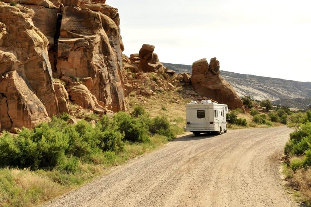 An RV driving on a dirt road with a large rock formation along the left side. 